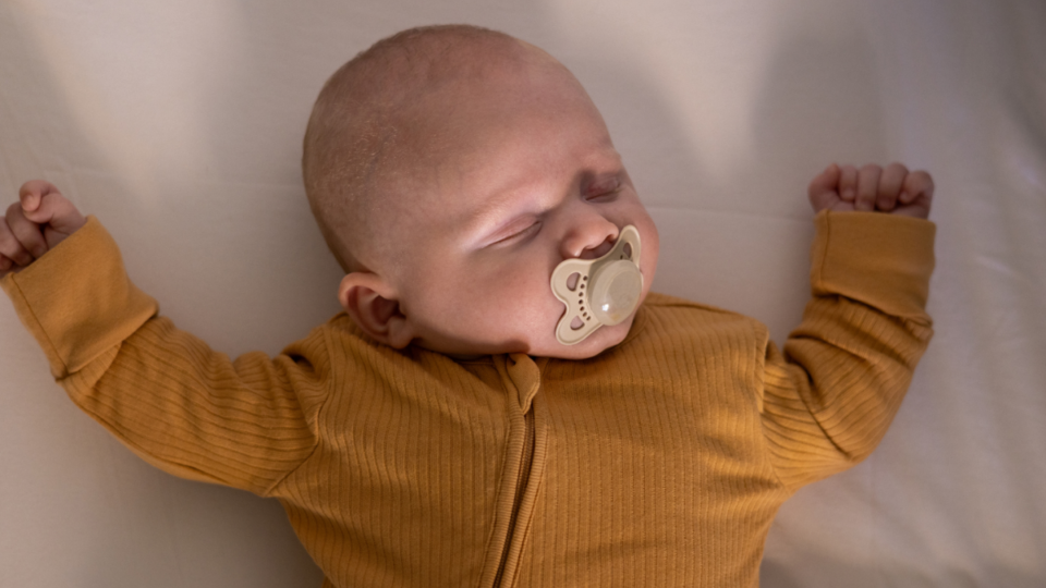 A baby is asleep in a safer sleep position, on their back with a clear cot. They have a dummy in their mouth.