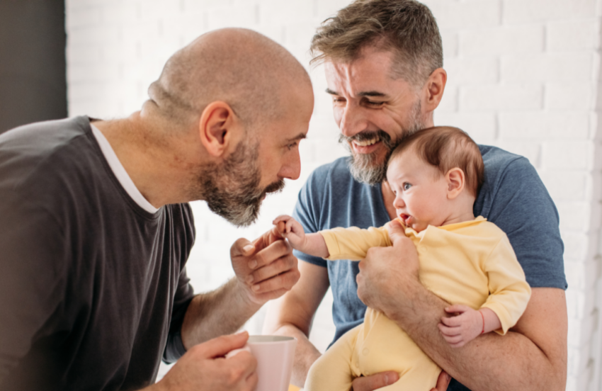 One man is holding a baby with the baby's head near his chin. The other man is holding the baby's hand in his, and putting his mouth to the baby's hand. Both men are smiling.