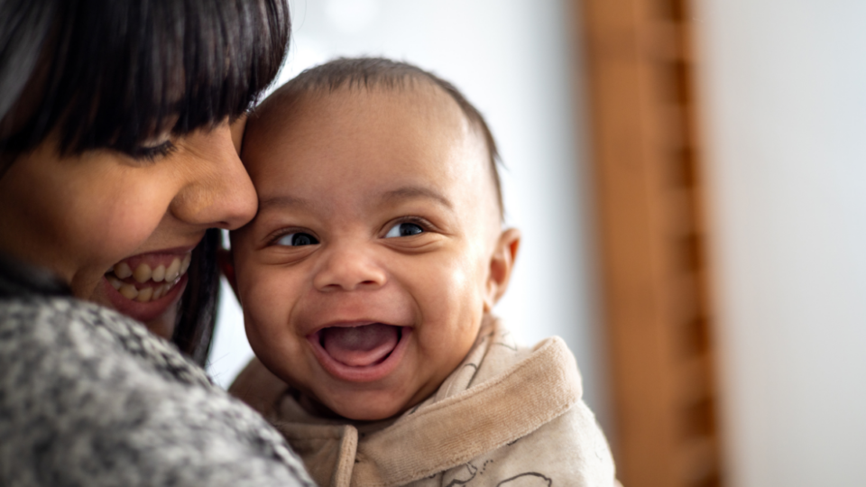 A baby is looking to their side with a huge, wide open grin. They have light to medium skin and the mum is looking down at them and smiling, with her face closely pressed to the baby.