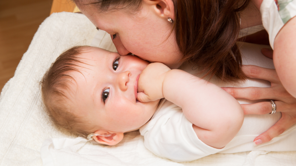 A baby is laid on a changing mat. Their mum is kissing their cheek, and the baby is looking directly at the camera, smiling with their hand in their mouth. They have blue eyes, light skin and hair and a hearing aid in their right ear.