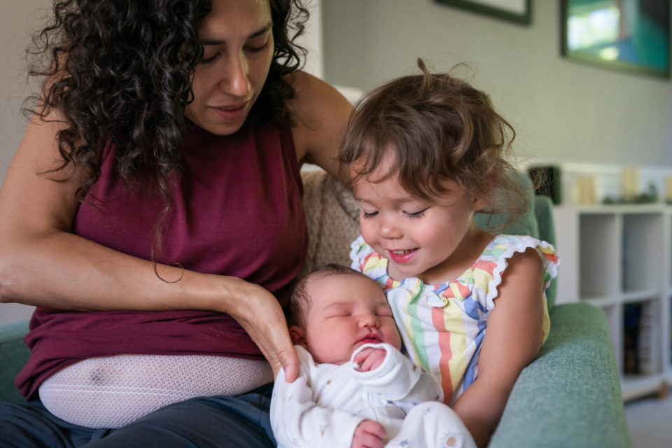 A woman sits on the sofa at home and helps her cute toddler daughter hold her baby brother for the first time. The big sister appears excited and proud.
