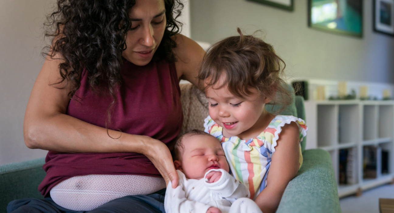 A woman sits on the sofa at home and helps her cute toddler daughter hold her baby brother for the first time. The big sister appears excited and proud.