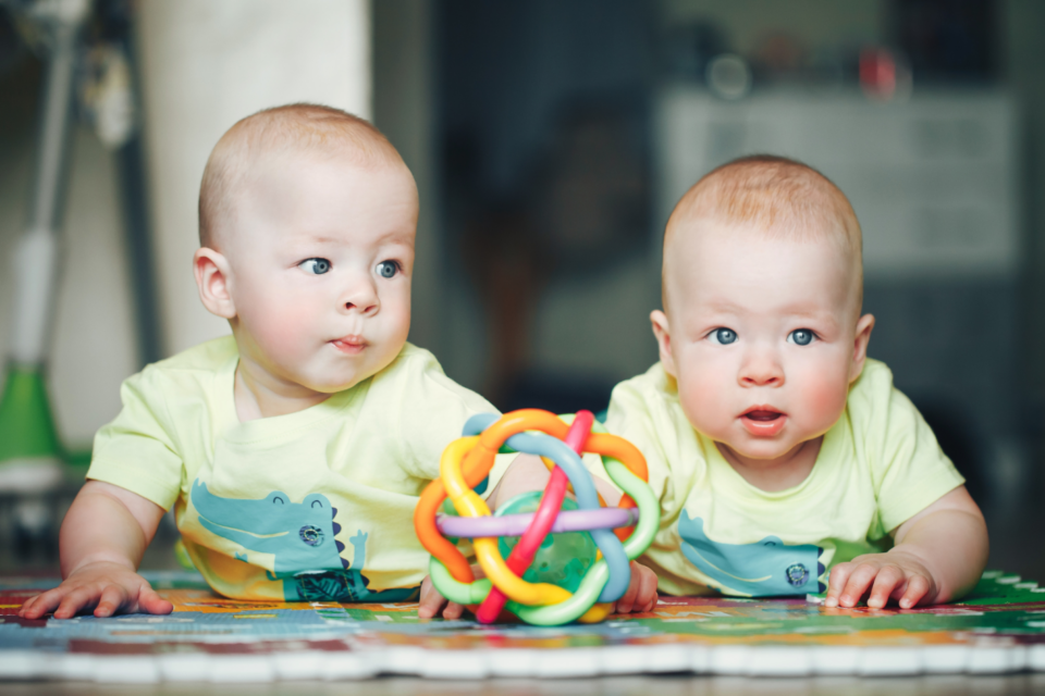Two young twin babies are having tummy time on a play mat with a toy. They have ginger hair and light skin with bright blue eyes.