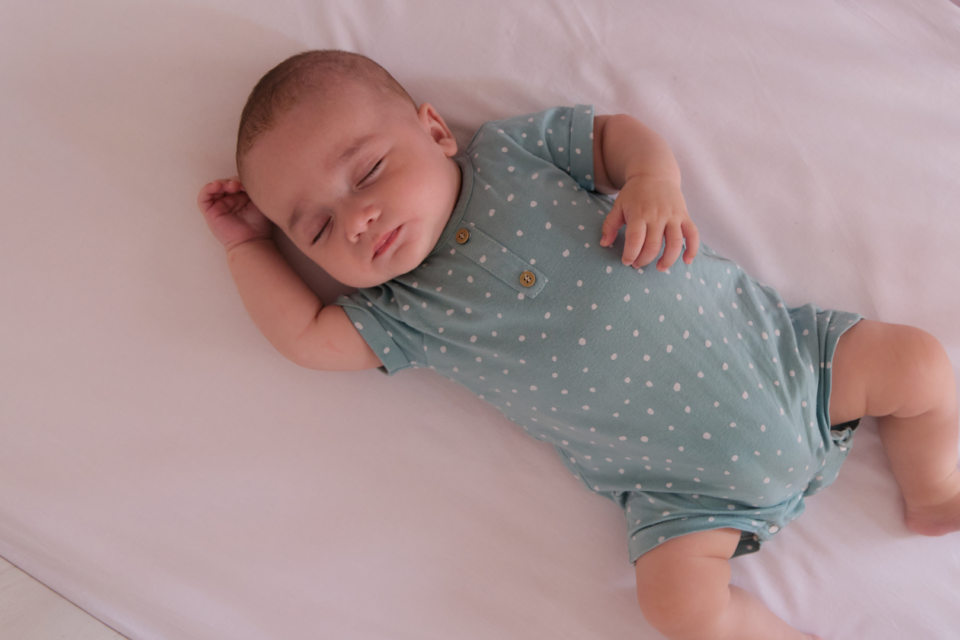 A baby sleeping safely on their back in an empty cot. They have one arm by their head, and another on their stomach. They are wearing a blue, polka-dot baby-grow.