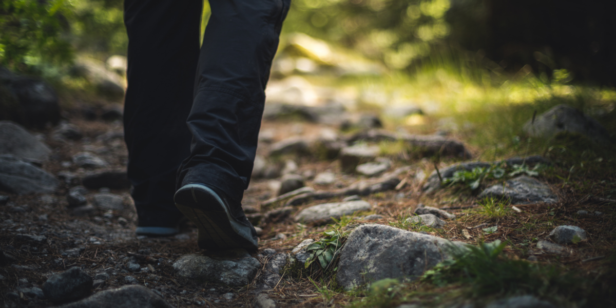 A man's legs and shoes are walking through grassy, rocky terrain.