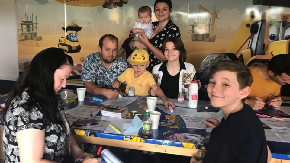 Family sitting at a table at a family day, doing colouring in and smiling at the camera.