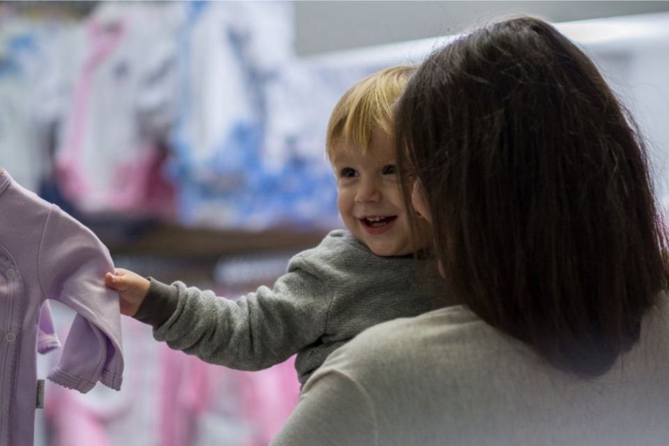 Mother in a shop holding her child who is smiling and holding onto an item of clothing on a hanger.