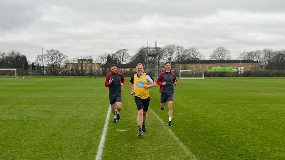 Karl running in a field wearing his yellow Lullaby Trust hoodie, with two footballers running behind him.