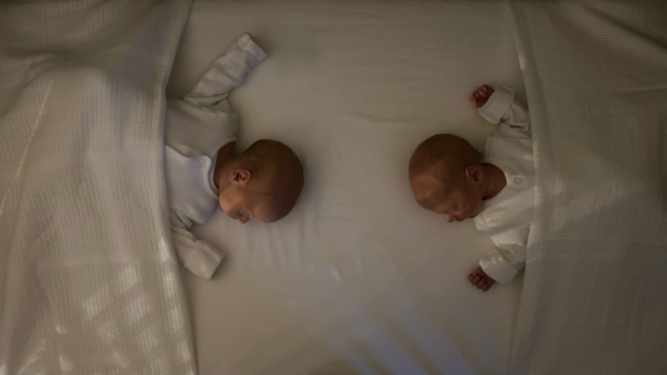 Twins co-bedding in a cot, feet to foot with blankets tucked in.