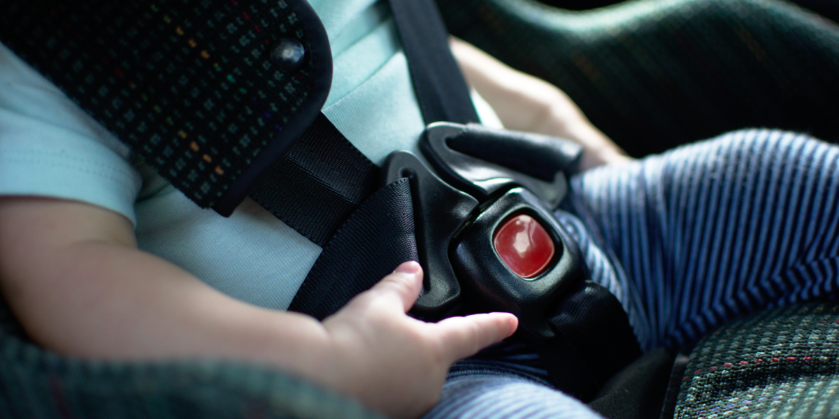 Close up of a baby in a car seat. You can see their mouth and the car seat buckled across their blue t-shirt. They are playing with the plastic around the buckle.