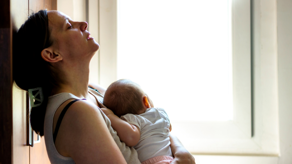 A tired mum is holding her baby and she is leaning up against a cabinet