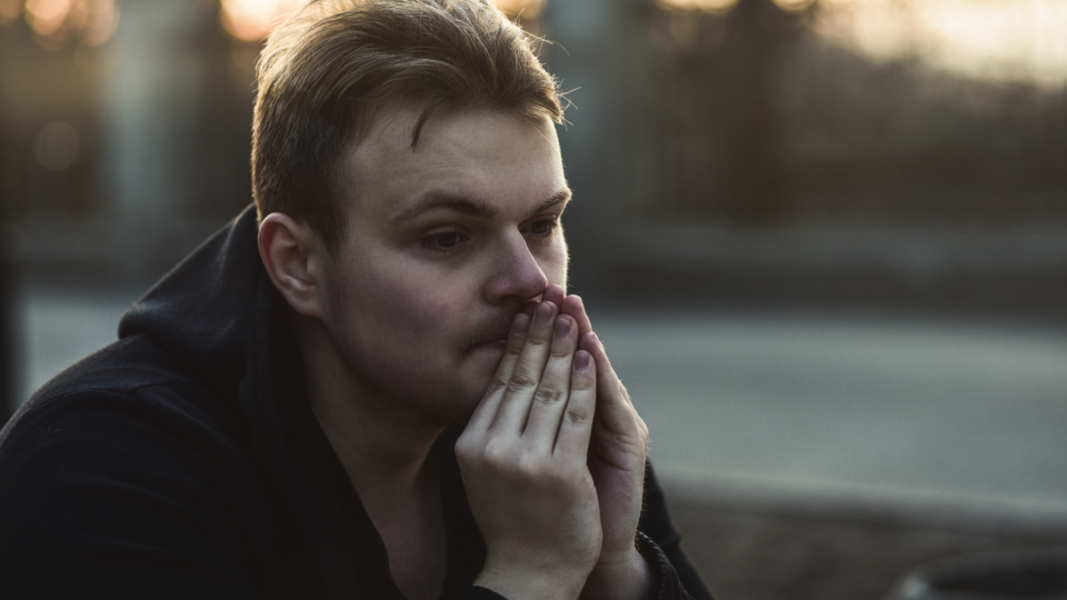 A man is looking into the distance with his hands up to his mouth. He looks deep in thought, and quite sad. The sun is setting behind him.