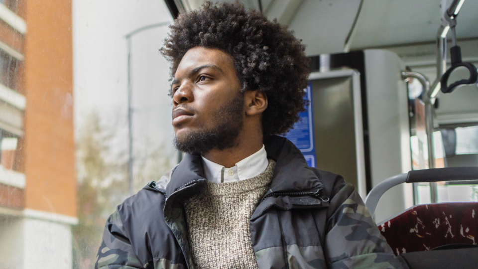 A man is looking out of the window, seemingly on a bus. He is wearing casual work-wear, and looks as though he's deep in thought, potentially sad.