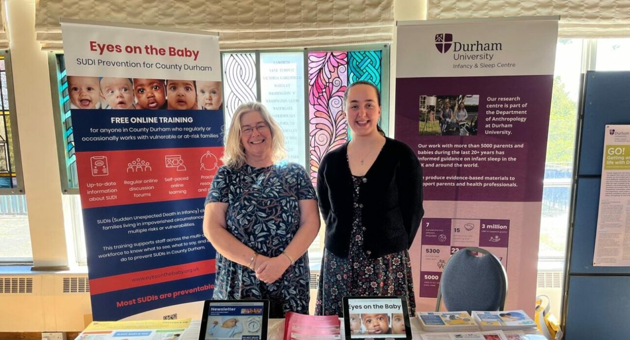 Two researchers are pictured next to one another, both women and both stood proudly. Behind them are pull up banners relating to SIDs and SUDI, and in front of them is a table full of useful information on their research projects.