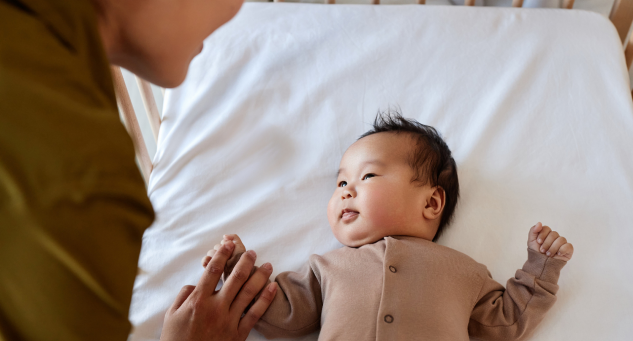 A baby is being put down to sleep in a cot. They are looking up at their parent.