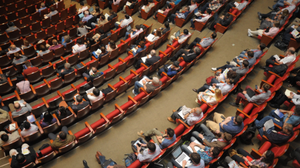 A shot from the top of an auditorium looking down at hundreds of people sat in seats at a conference.