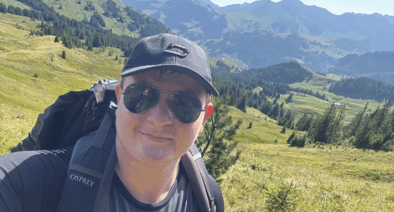Matt wearing a cap and sunglasses, with a backpack on his back. He is taking a selfie in front of a vast natural landscape.