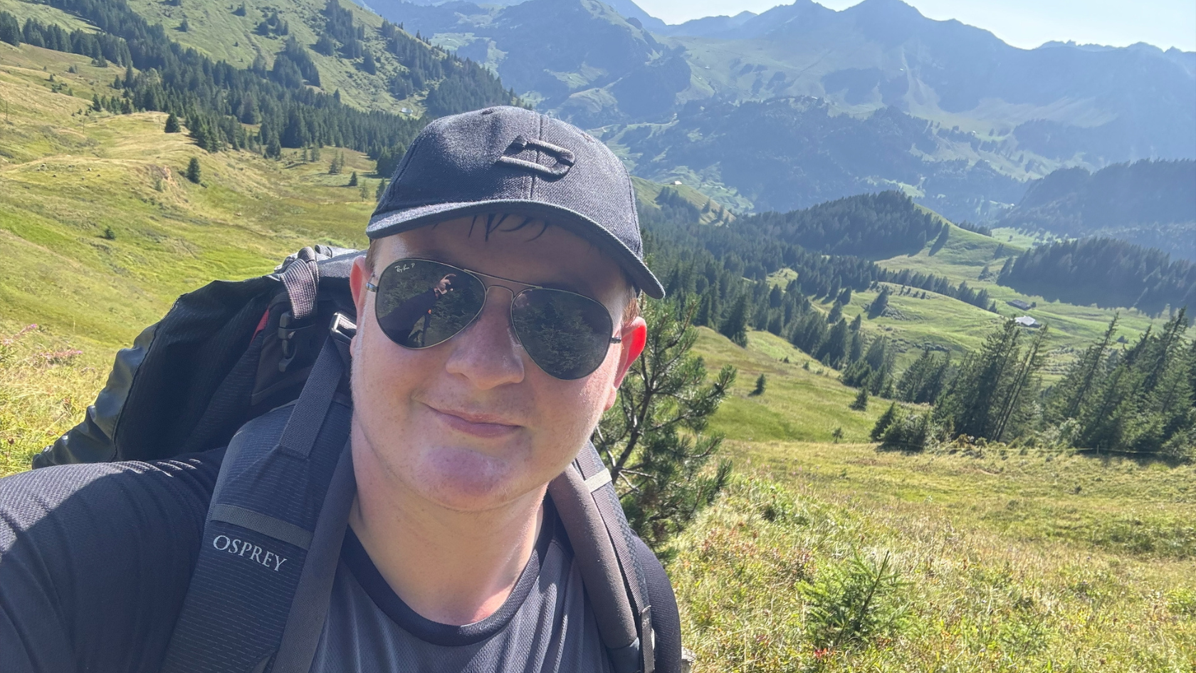 Matt wearing a cap and sunglasses, with a backpack on his back. He is taking a selfie in front of a vast natural landscape.