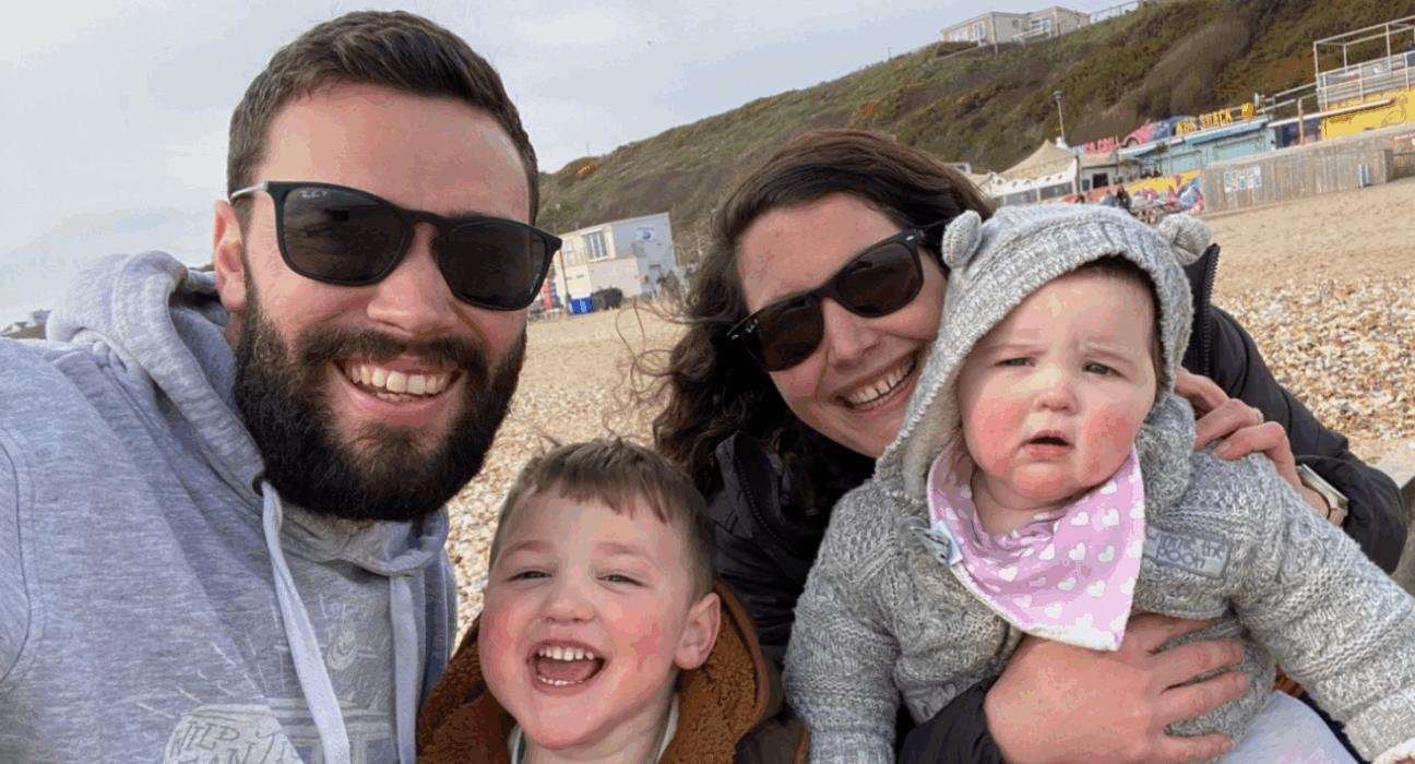 Beth and Callum with three-year-old Rory and eight-month-old Poppy. They are taking a selfie on a pebbly beach.