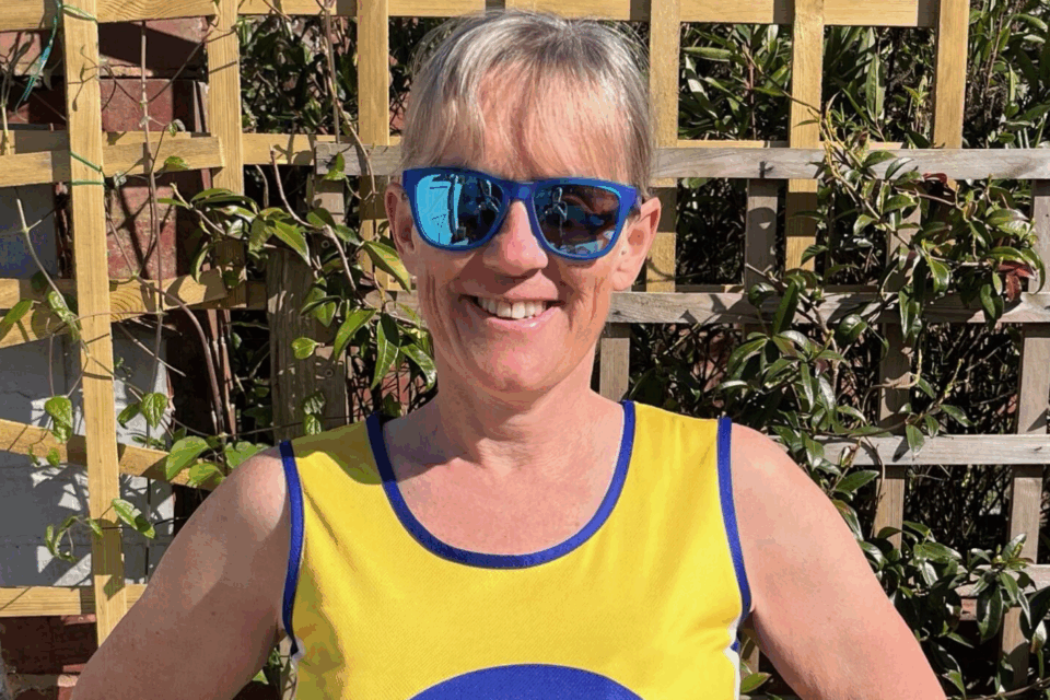 Clare wearing sunglasses and her Lullaby Trust vest, standing outdoors smiling at the camera.