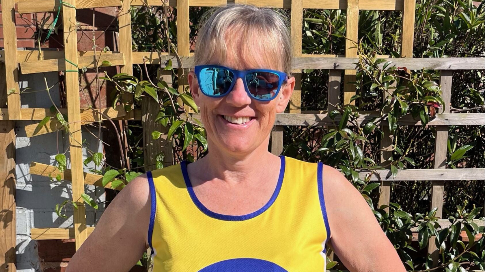 Clare wearing sunglasses and her Lullaby Trust vest, standing outdoors smiling at the camera.