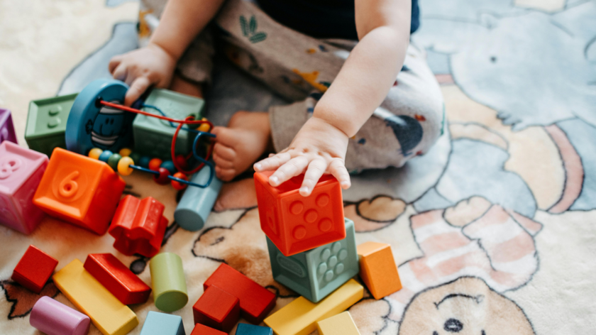 A close up of a baby's hand playing with some blocks. The baby is sitting, playing with plastic and wooden colourful blocks on a mat.