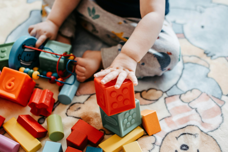 A close up of a baby's hand playing with some blocks. The baby is sitting, playing with plastic and wooden colourful blocks on a mat.