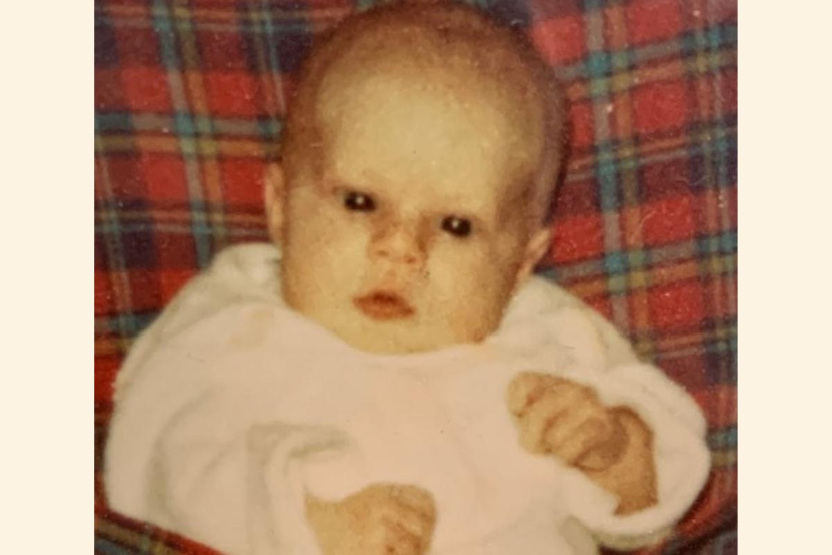Baby Gayle wearing a white top, sitting on a tartan seat.
