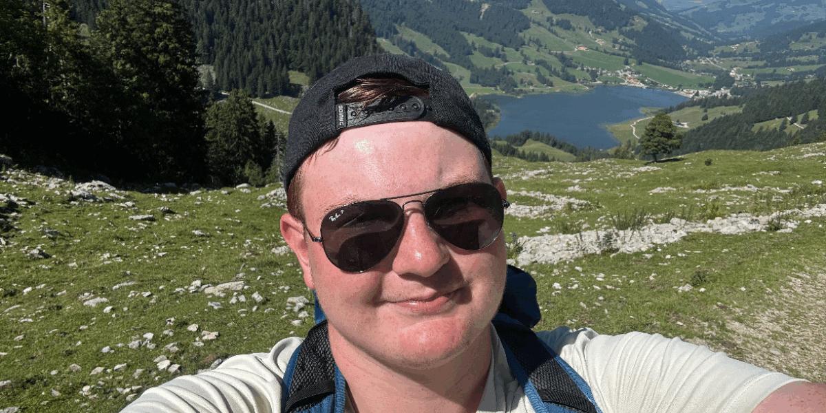 Matt wearing sunglasses and a backwards cap, taking a selfie in front of a green landscape with mountains.