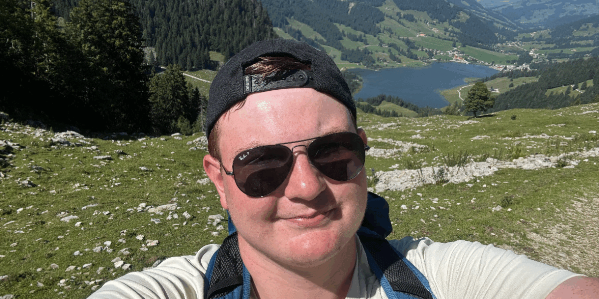 Matt wearing sunglasses and a backwards cap, taking a selfie in front of a green landscape with mountains.