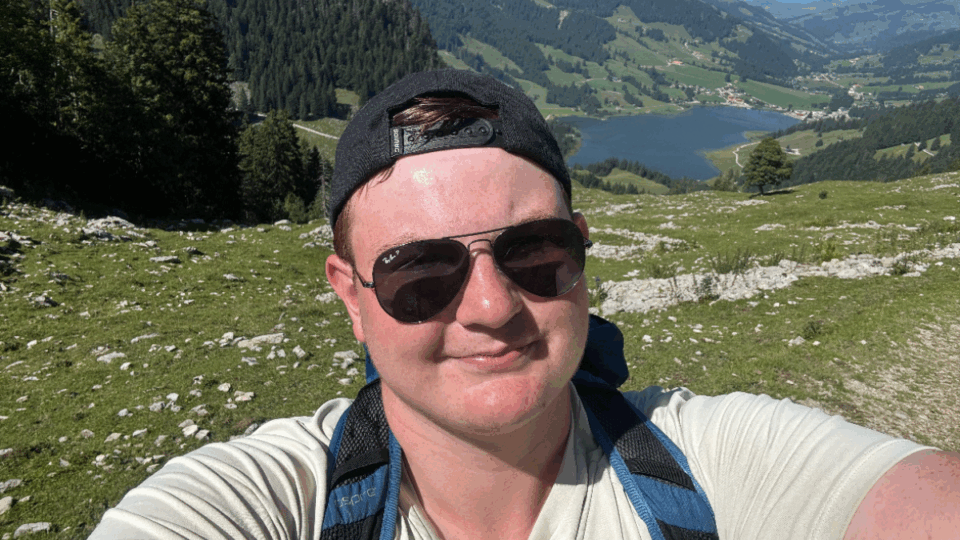 Matt wearing sunglasses and a backwards cap, taking a selfie in front of a green landscape with mountains.