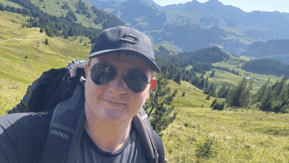 Matt wearing a cap and sunglasses, with a backpack on his back. He is taking a selfie in front of a vast natural landscape.