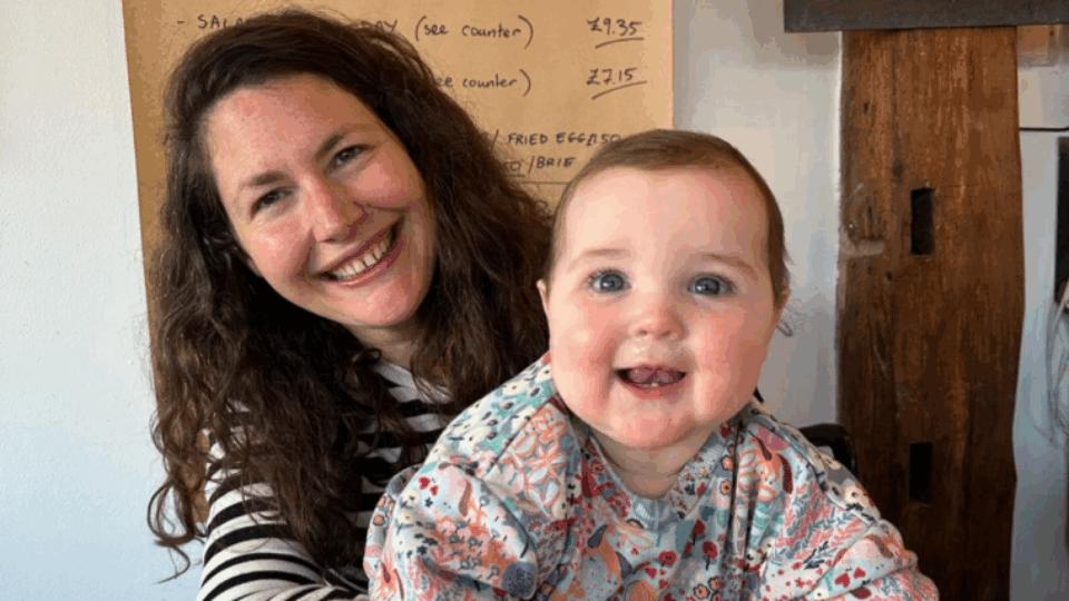 Beth sitting in a cafe wearing a stripy top, holding Poppy. Both are smiling at the camera.