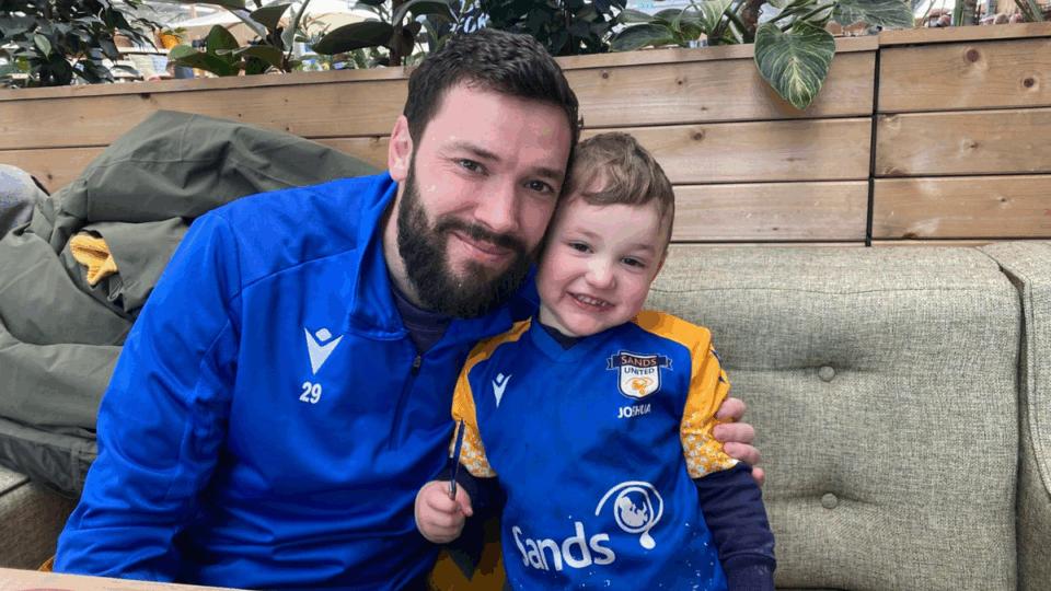 Callum and Liam sitting together, both wearing blue football tops, smiling at the camera.