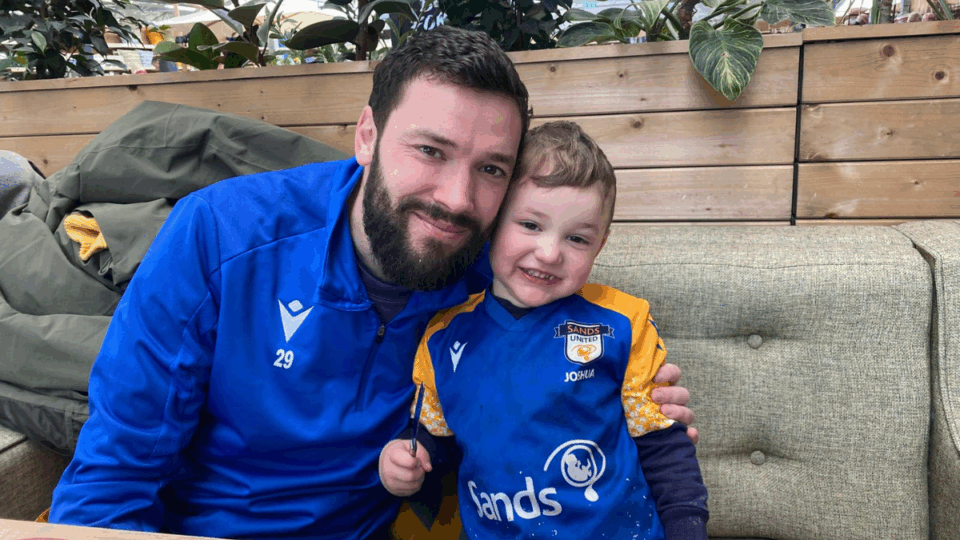 Callum and Liam sitting together, both wearing blue football tops, smiling at the camera.
