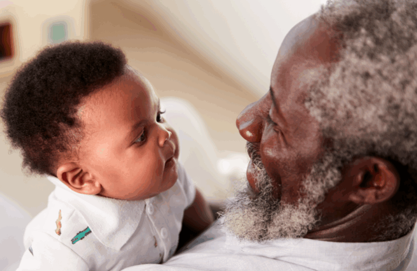 A baby is looking at his grandparent and their faces are close together. The granddad has dark skin and hints of grey in their hair and bears, and the baby is wearing a baby grow.
