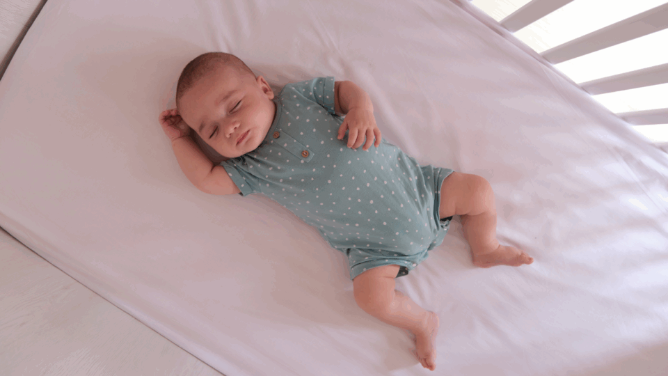Baby sleeping in a cot on their back. They have a light sleep suit on and the light is coming through the bars of the cot. They have light skin.