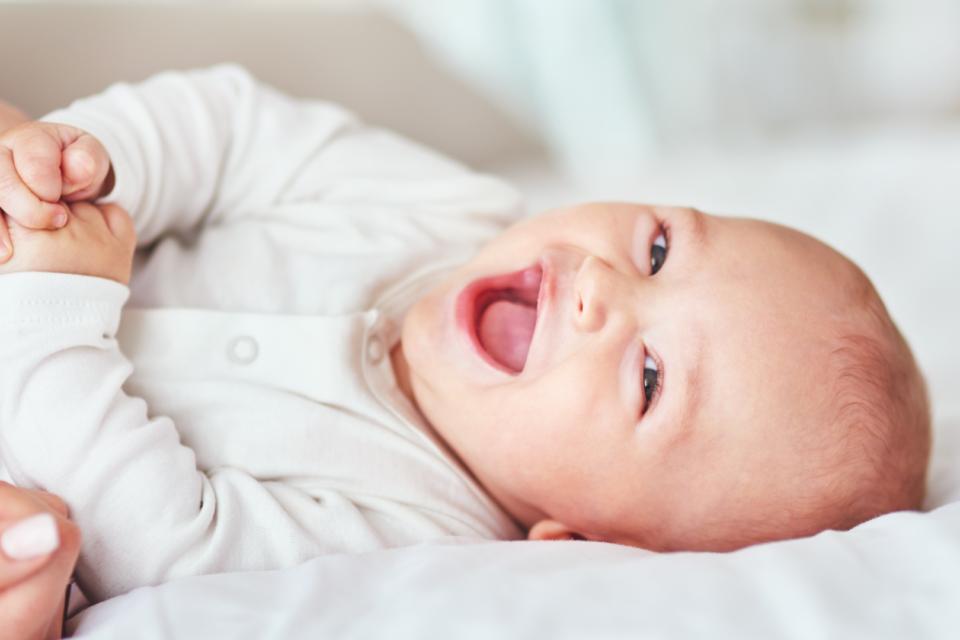 Baby is laying down smiling and giggling, grasping their hands together. They have light skin and wear a white babygrow.