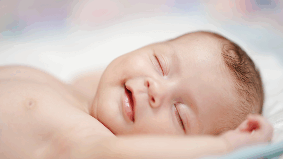 A baby is sleeping and smiling in their sleep. Their arms are either side of their head.