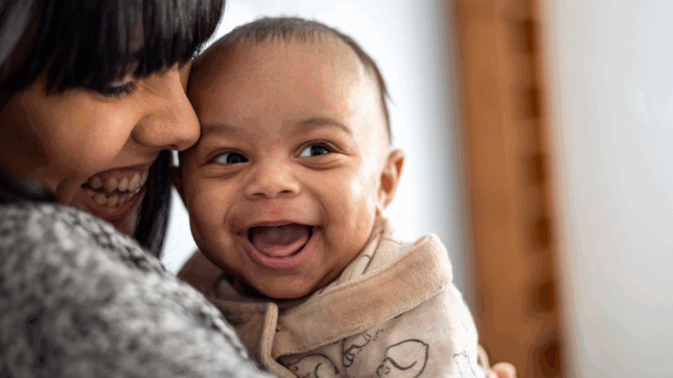 A baby is looking to their side with a huge, wide open grin. They have light to medium skin and the mum is looking down at them and smiling, with her face closely pressed to the baby.