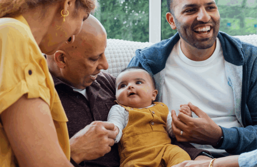 A family are gathered around their baby. They are all smiling and looking at the baby and the baby is sat in between two of the men.