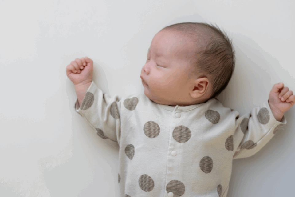 A baby is sleeping in on their back, with their head tilted to the side. They have dark hair, light skin and wear a polkadot baby grow. Their arms are rested either side of their head.