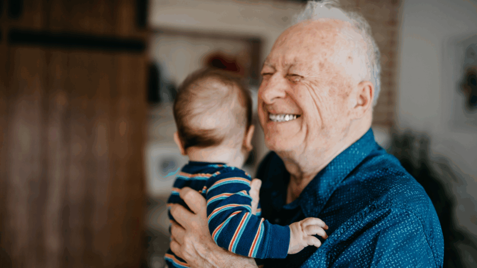Grandad standing up smiling, holding a baby who is looking away.