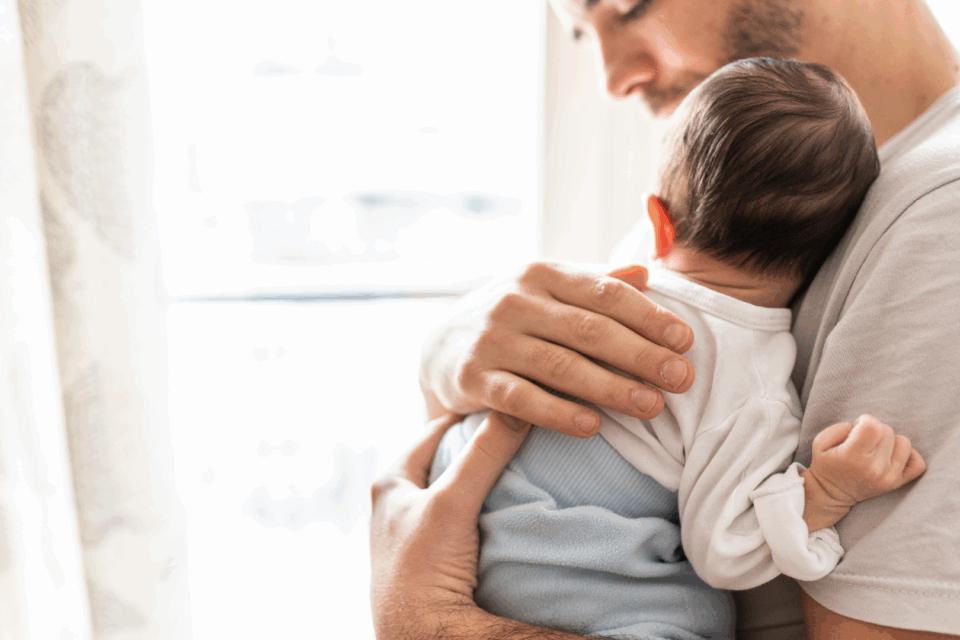 A dad is holding their baby on their chest, with his hand on the baby's back.