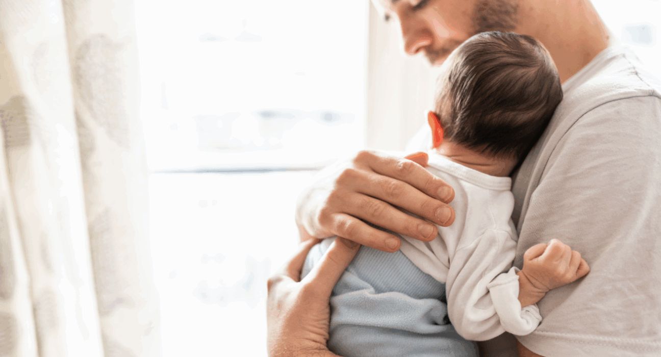 A dad is holding their baby on their chest, with his hand on the baby's back.