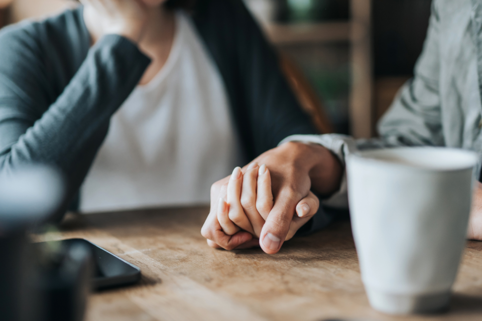 Two people are holding hands supportively. There is a phone and mug on the table.