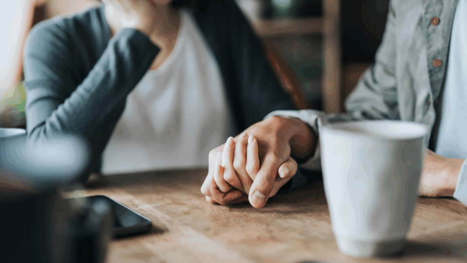 Two people are holding hands supportively. There is a phone and mug on the table.