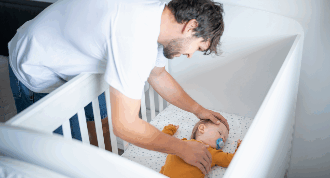 A baby is sleeping in a safer sleep position in a cot. The dad is holding a hand on his chest and head. The baby has a dummy.