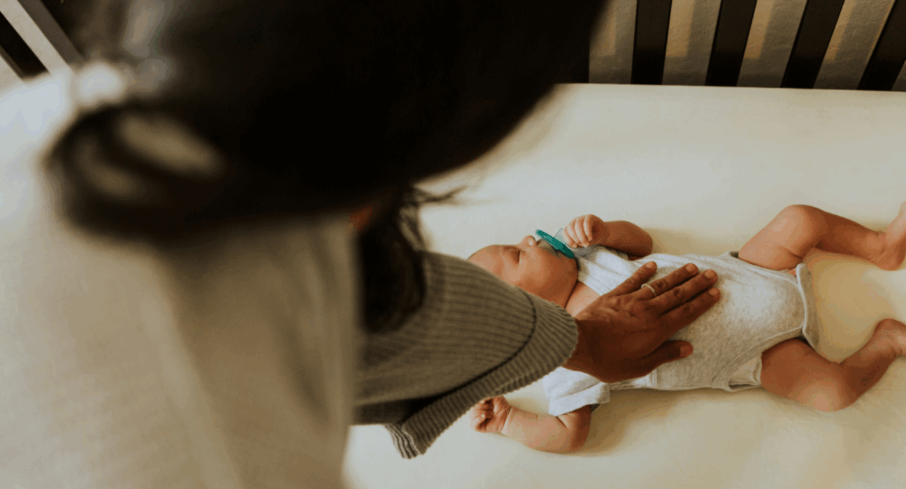 A baby is sleeping on their back in their cot with a dummy in their mouth. They have a medium skin tone and their mum has her hand on their chest.