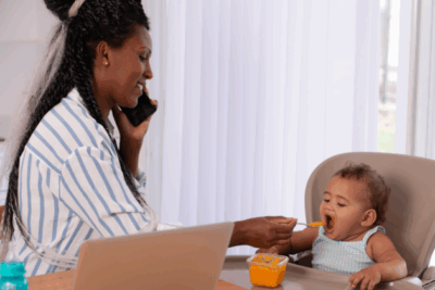 A mum is feeding her baby in their highchair, whilst on the phone with the other hand. She is smiling.